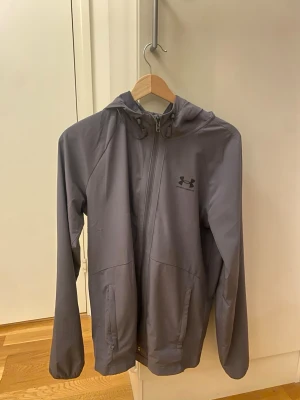 Grå vindjacka från Under Armour - Snygg grå vindjacka från Under Armour med huva och dragkedja framtill. Jackan har två sidofickor och diskret logga på bröstet. Perfekt för blåsiga dagar och sportiga outfits. Tillverkad i lätt och smidigt material.