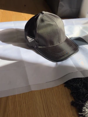 Svart keps från Prada i nylon - Snygg svart keps från Prada i glansigt nylonmaterial. Klassisk böjd skärm och ikonisk metallogo på sidan. Perfekt accessoar för en trendig streetwear-look.