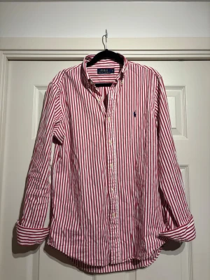 Rödvit randig skjorta Polo Ralph Lauren - Klassisk skjorta från Polo Ralph Lauren i rödvitt randigt mönster. Skjortan har lång ärm, knappar framtill och den ikoniska broderade loggan på bröstet. Grym till sommaren!
