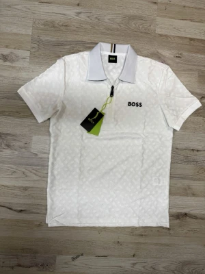 Vit BOSS t-shirt med dragkedja - Snygg vit t-shirt från BOSS med subtilt mönster och svart BOSS-logga på bröstet. T-shirten har korta ärmar, klassisk krage och en liten dragkedja framtill. Perfekt för en stilren och modern look.