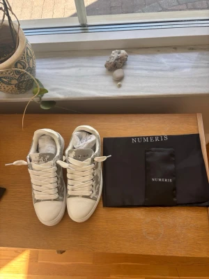 Numeris - Snygga vita sneakers från Numeris med grått kamouflagemönster på sidorna och vita detaljer. Skorna har tjock, räfflad sula och breda vita skosnören. Tillverkade i textil och syntetmaterial. Perfekta för dig som vill sticka ut med en unik look.