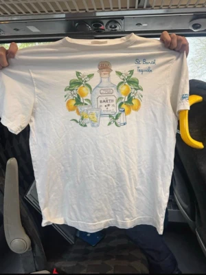 Vit St Barth Tequila t-shirt - Vit t-shirt från St Barth med färgglad tequila- och citronprint på framsidan. Broderad text 'St Barth Tequila' i blått på bröstet. Klassisk passform och korta ärmar, perfekt för en avslappnad look. Den är knappt anvönd med är skrynklig eftersom den legat dåligt vikt