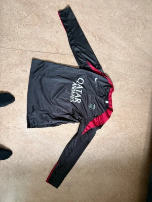 Svart PSG långärmad träningströja Nike - Säljer en svart långärmad träningströja från Nike med PSG-logga och Qatar Airways-tryck på bröstet. Tröjan har röda detaljer längs axlar och ärmar, samt rund hals. Perfekt för fotbollsträning eller sportaktiviteter.