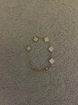 Guldarmband med vita fyrklöver - Snyggt armband i guldton med fem vita fyrklöverformade hängen. Hängena har en pärlemorskimrande yta och är infattade i guldfärgad metall. Kedjan är tunn och elegant, perfekt för att ge en lyxig touch till din stil.