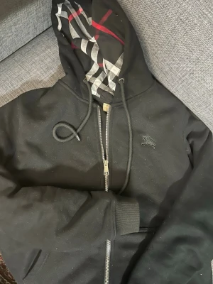 Svart hoodie från Burberry - Säljer en svart hoodie från Burberry med dragkedja och klassiskt rutigt foder i huvan. Hoodien har snörning i huvan, ribbade muddar och diskret Burberry-logga på bröstet. Materialet är mjukt och bekvämt, perfekt för en avslappnad stil. Köpte den förra veckan, säljer pga fel storlek!