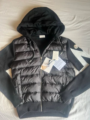 Svart Moncler hybridjacka med huva - Snygg svart hybridjacka från Moncler med vadderad framsida, stickade ärmar och huva. Dragkedja framtill, två fickor med knapp och ikonisk Moncler-logga på ärmen. Materialmix av dun och ull ger både värme och stil. Perfekt för dig som vill ha en exklusiv streetwear-look.