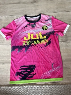 Rosa sporttröja JUL Toto & Nineta - Säljer en rosa sporttröja från JUL med tryck i gult och svart, samt texten 'Toto & Nineta' på framsidan. Tröjan har korta ärmar, svart krage och vita detaljer vid ärmsluten samt en grön rand längst ner. Perfekt för träning eller match.