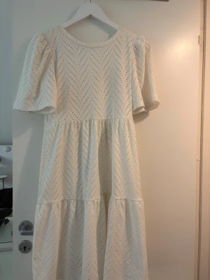 Vit klänning med broderade detaljer - Superfin vit klänning, i knälängd med broderade hålmönster och puffiga korta ärmar. Klänningen har en luftig passform och är perfekt för varma dagar. Materialet känns mjukt och andas, och den har en romantisk vibe med volang i kjolen.