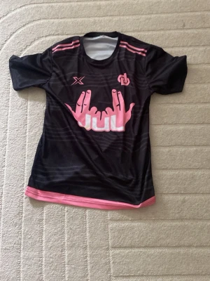 Svart sporttröja med rosa detaljer - Snygg svart sporttröja med rosa detaljer och tryck framtill. Tröjan har korta ärmar, rund halsringning och är gjord i ett lätt, snabbtorkande material som passar perfekt till träning. Rosa grafiskt tryck och loggor på bröstet ger en cool look.