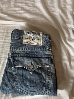True Religion blå jeans - Säljer ett par klassiska blå jeans från True Religion med ikoniska kontrastsömmar. W33. Jeansen har bakfickor med lock och knapp samt en tydlig logga på bakfickan. Tillverkade i slitstark denim med snygg tvätt. Säljer eftersom jag bytt stil och inte använder dom mer och för att jag behöver pengar till sommaren.