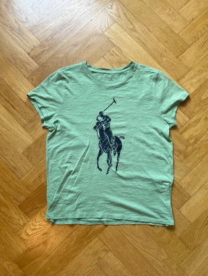 Grön t-shirt från Polo Ralph Lauren - Snygg ljusgrön t-shirt från Polo Ralph Lauren med det klassiska stora polospel-trycket i svart på bröstet. T-shirten har rund halsringning och korta ärmar. Tillverkad i mjuk bomull som känns skön mot huden. Perfekt för en avslappnad och stilren look.