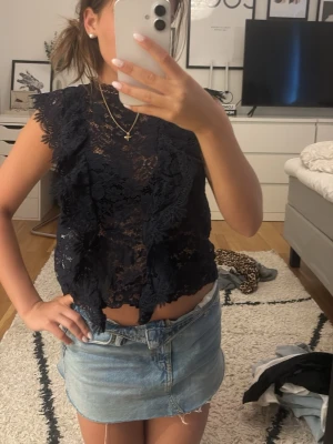 Svart spetsblus med volangdetaljer - Säljer en svart, ärmlös blus i spets med volangdetaljer framtill och längs axlarna. Toppen har hög hals och är transparent, vilket ger en trendig och elegant look. Perfekt att styla med jeans eller kjol för en cool outfit.
