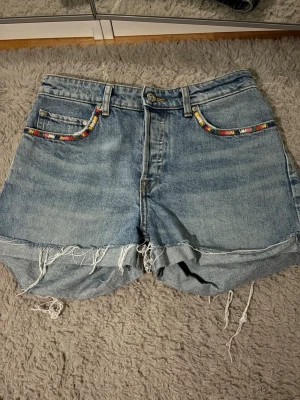 Blå denimshorts med broderade detaljer - Säljer ett par blå denimshorts med råa, fransiga kanter och broderade detaljer i färg vid fickorna fram.