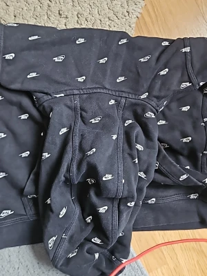 Svart Nike hoodie med loggor XL - Svart hoodie från Nike i storlek XL, täckt med små vita Nike-loggor över hela plagget. Klassisk huva och magficka framtill. Tillverkad i mjukt material som passar perfekt för en avslappnad streetwear-look.
