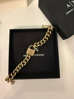 Guldfärgat armband Armani Exchange - Snyggt guldfärgat armband från Armani Exchange med grov kedja och ett hängande lås med A|X-logga. Armbandet har en cool och modern design som sticker ut och passar perfekt till dig som gillar statement-smycken. Levereras i original ask.