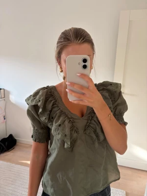 Olivgrön blus med volanger från Vila - Trendig olivgrön blus från Vila med broderade volanger längs v-ringningen och axlarna. Blusen har korta puffärmar, den är i storlek 34!💕💕