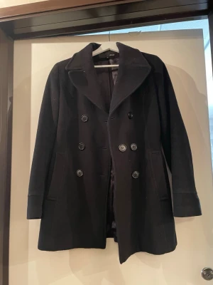 Svart dubbelknäppt  - Stilren svart kappa från HENNES. Kappan har två fickor framtill och långa ärmar med breda muddar. Perfekt för kalla dagar när du vill ha en klassisk look. Funkar för höst, vinter och vår. 