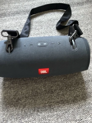 JBL Xtreme 2 trådlös högtalare - Säljer en JBL Xtreme 2 trådlös högtalare i mörkgrå färg med robust tygmaterial och bärrem. Högtalaren har tydliga kontrollknappar på ovansidan och JBL-logga framtill. Laddare och strömkabel medföljer. Perfekt för att ta med sig musiken överallt.