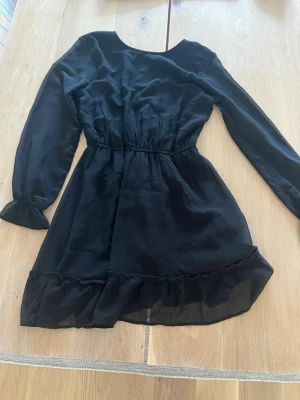 Svart volangklänning från Kappahl - Snygg svart klänning från Kappahl med långa ärmar och volangdetaljer nertill. Klänningen har markerad midja och är tillverkad i ett lätt, flowy material som ger en skön känsla.
