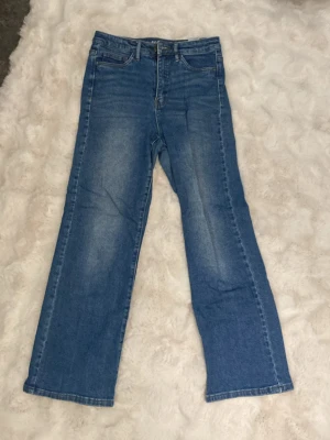 Blå raka jeans med hög midja - Säljer ett par klassiska blå jeans med raka ben och hög midja. Jeansen har fem fickor, dragkedja och knapp framtill samt snygga slitningar på låren för en avslappnad look. Perfekta till en casual outfit och tillverkade i denim.