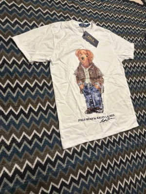 Vit Polo Bear t-shirt från Ralph Lauren - Vit t-shirt från Ralph Lauren med ikoniska Polo Bear-trycket framtill. Klassisk rund hals och korta ärmar. Tillverkad i mjuk bomull för skön känsla och stilren look. Perfekt för dig som gillar grisch och vill ha en snygg stil den är oanvänd och är i storlek s