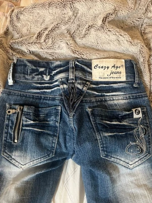 Coola jeans från Crazy Age - Säljer ett par blå bootcut jeans från Crazy Age med tribal-mönster i svart på benen och bakfickan. Jeansen har svarta detaljer med rinestones, kontrastsömmar och en cool dragkedja på ena bakfickan. Materialet är klassisk denim i bomull.                       Stl W22/xxs        Lågmidjade          Har sytt upp så passar någon som är 154-159 ungefär 💕.         Går att sänka pris vid snabb affär /pruta och förhandla 💕