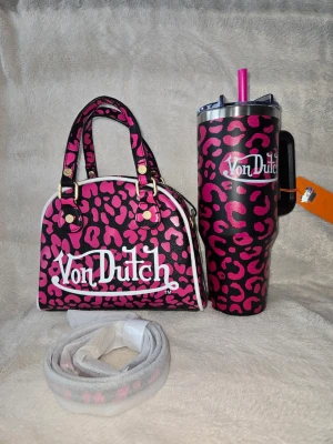 Von Dutch rosa leopard handväska och matchande termosmugg  - Cool handväska från Von Dutch i svart och rosa leopardmönster med vit logga framtill. Väskan har två handtag med guldfärgade detaljer och vit kant runtom. Perfekt statement piece för dig som gillar att sticka ut och en matchande termosmugg