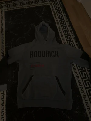 Grå HOODRICH hoodie med tryck - Snygg grå hoodie från HOODRICH med svart tryck över bröstet och detaljer i svart och rött. Hoodien har en stor huva och magficka framtill. Perfekt för en avslappnad streetwear-look.