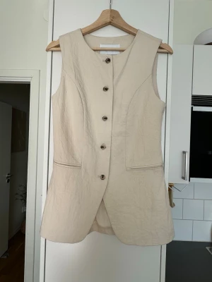 Beige lång väst med knappar - Stilren beige väst i längre modell med fem bruna knappar framtill och två fickor. Västen har en rak passform och är ärmlös, perfekt för lager-på-lager. Materialet ser ut att vara linne eller bomull för en naturlig känsla.