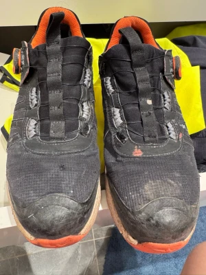 Svarta sneakers med orange detaljer - Svarta sneakers med orange insida och detaljer. Skorna har en unik snörning med breda band och gråa accenter vid snörhålen. Ovandelen är i textil och sulan är vit med orange framtill. Perfekta för en sportig och avslappnad stil.