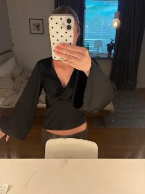 Svart omlottopp med vida ärmar - Snygg svart omlottopp med v-ringning och vida långärmar. Toppen har en croppad passform och knytdetalj framtill. Perfekt för att lyfta vilken outfit som helst.