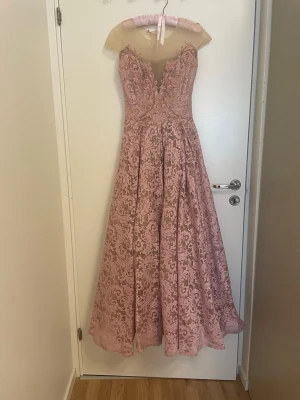 Rosa långklänning  - Elegant rosa långklänning med v-ringad spets upptill och genombrutna detaljer. Klänningen har ett vackert blommigt mönster och en lätt utsvängd kjol. Perfekt för bal eller festliga tillfällen. Axelpartiet är i transparent mesh och klänningen har dragkedja bak.