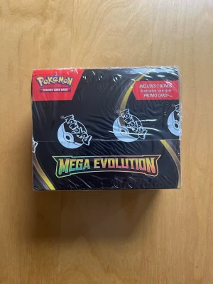Pokémon Mega Evolution boosterbox - Säljer en förseglad Pokémon Mega Evolution boosterbox med officiell plastförsegling. Lådan är svart med färgglad text och innehåller samlarkort samt en bonus illustration rare-style promo card. Perfekt för dig som vill utöka din samling eller spela Pokémon TCG. (obs ej äkta) 