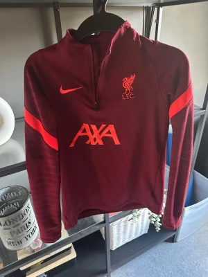 Vinröd Liverpool träningströja Nike - Snygg vinröd långärmad träningströja från Nike med Liverpool FC-logga och AXA-tryck på bröstet. Tröjan har halv dragkedja framtill och röda detaljer på ärmarna. Perfekt för fotbollsträning eller som sportig vardagsstil.