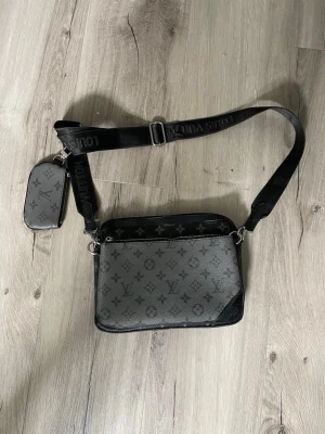 axelväska monogram svart - Snygg axelväska i svart och grått med klassiskt monogrammönster. Väskan har justerbar axelrem med logotyp och en mindre avtagbar ficka. Tillverkad i canvas med detaljer i läder och metall.axelväska monogram svart
