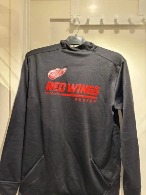 Autentisk Detroit Red Wings hoodie | JR XL  - Snygg svart långärmad hockeytröja från Fanatics med Detroit Red Wings-logga och text i rött framtill. Tröjan har hög krage och två fickor framtill. Perfekt för dig som gillar NHL och vill ha en sportig look. Köpt via NhL:s officiella hemsida och har alla autenticitetsgrejer.