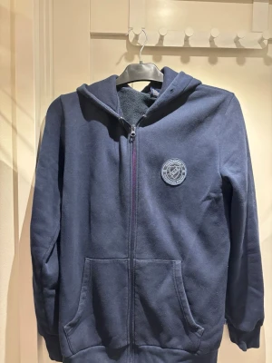 Blå Djurgården zip hoodie med dragkedja | strl S - Mörkblå hoodie med dragkedja från Djurgårdens IF. Har klubbens emblem broderat på bröstet och två stora fickor framtill. Utrustad med huva och tillverkad i mjukt bomullsmaterial. Perfekt för dig som vill visa ditt stöd för DIF.Köpt i Djurgårdsbutiken!
