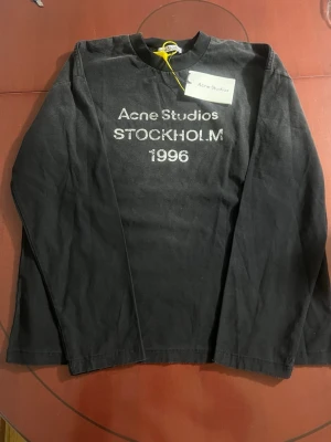 Svart långärmad topp Acne Studios - Svart långärmad topp från Acne Studios med tryck 'Acne Studios STOCKHOLM 1996' på bröstet. Klassisk passform och rund halsringning. Tillverkad i mjuk bomull, perfekt för en clean och stilren look. OBS! Original storlek är S men den är mer baggy i storlek å passar därmed M