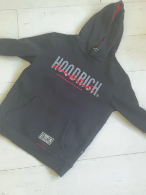 Svart HOODRICH hoodie med tryck - Svart hoodie från Hoodrich med stor logga och röd detalj på bröstet. Klassisk känguruficka framtill och huva med röd insida. Perfekt för en streetwear-look och riktigt skön att bära.