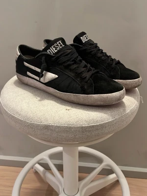 Svarta Diesel sneakers med vita detaljer - Snygga svarta sneakers från Diesel med vita detaljer och logga på plösen. Skorna har lågt skaft, svarta skosnören och en röd sula med vintage-look. Materialmix av mocka och textil ger en cool och urban känsla. Stor i storlek och köpta för 2400