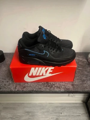 Nike Air Max 90 svart/blå sneakers - Snygga Nike Air Max 90 sneakers i svart med blå detaljer och logga. Skorna har en klassisk låg siluett, mesh och läder på ovandelen samt den ikoniska Air Max-sulan för extra komfort. Perfekta för dig som vill ha en stilren och sportig look.