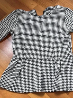 Svartvit peplumblus med mönster - Snygg svartvit peplumblus med klassiskt hundtandsmönster. Blusen har lång ärm och en högre halsringning. Materialet känns mjukt och stretchigt, perfekt för en trendig look. Passar dig som gillar stilrena och tidlösa plagg.