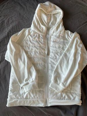 Vit cardigan - Vit hoodiejacka med quiltade partier och hel dragkedja framtill. Jackan har huva, långa ärmar och ribbade muddar. Perfekt för lager-på-lager och en clean look.