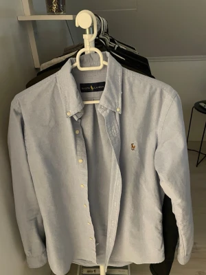 Ljusblå skjorta från Ralph Lauren - Klassisk ljusblå skjorta från Ralph Lauren med button-down krage och broderad logga på bröstet. Skjortan har långa ärmar och knäppning framtill. Perfekt för en stilren och avslappnad look. Passar även M