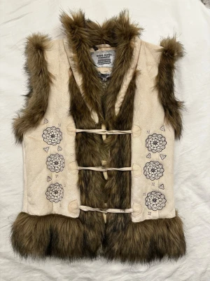 Pälsväst/Teddyväst - Miss Posh - Snygg beige pälsväst från Miss Posh med bruna fuskpälskanter och detaljerade blommönster på framsidan. Västen har tre knytband framtill och är fodrad med mjuk fuskpäls på insidan. Perfekt för att lyfta din outfit och ser bra ut över tröja eller hoodie.