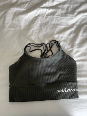 Grön sport-bh från xshapes - Snygg grön sport-bh från xshapes med ribbat tyg och brottarrygg med flätade och tunna axelband. Perfekt för träning eller yoga. Diskret logga broderad framtill. Materialet är stretchigt och bekvämt.