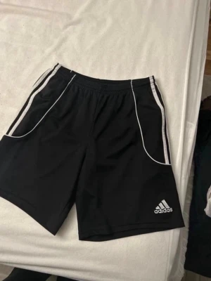 Svarta Adidas climalite shorts med vita ränder - Svarta sportiga shorts från Adidas med vita ränder längs sidorna och vit piping vid fickorna. Klassisk logga på ena benet och elastisk midja för skön passform. Perfekta för träning eller chill.