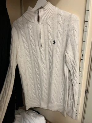 Vit kabelstickad tröja från Ralph Lauren - Kabelstickad vit tröja från Ralph Lauren med halv dragkedja och hög krage. Tröjan har långa ärmar, ribbstickade muddar och den klassiska broderade loggan på bröstet. Perfekt för dig som gillar stilren och tidlös design.