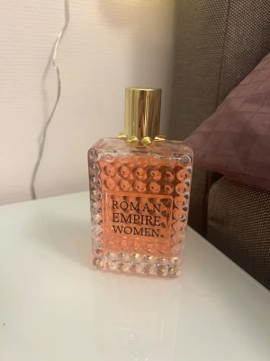 Roman Empire Women parfym - Säljer en snygg parfymflaska från Roman Empire Women. Flaskan är rektangulär med bubblig struktur, genomskinlig med rosa vätska och har ett lyxigt guldigt lock. Perfekt som en statement-doft på hyllan.