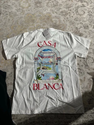 Vit Casablanca t-shirt  - Säljer en vit t-shirt från Casablanca med färgglatt tryck av ett fönster och texten 'Casa Blanca' på ryggen. T-shirten har korta ärmar och är gjord i mjuk bomull. Perfekt för dig som gillar stilrena plagg med unika detaljer.